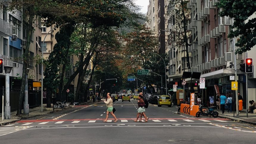 Pessoas atravessam uma rua molhada em Copacabana, no Rio de Janeiro, sob chuva, cercadas por edifícios urbanos.