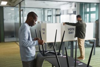 Duas pessoas preparam cabines de votação em um ambiente interno para o dia da eleição.