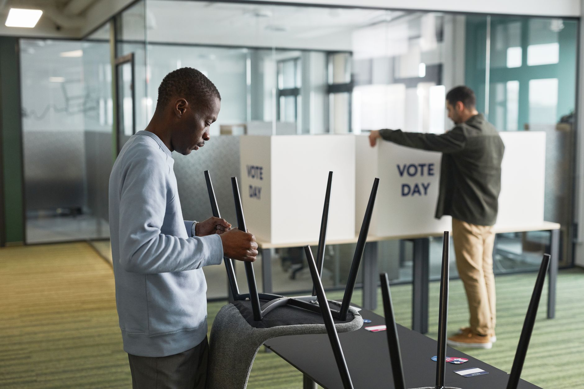 Duas pessoas preparam cabines de votação em um ambiente interno para o dia da eleição.