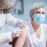 Paciente idosa recebe uma vacina no braço enquanto é atendida por uma profissional de saúde usando equipamentos de proteção.