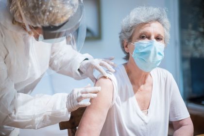 Paciente idosa recebe uma vacina no braço enquanto é atendida por uma profissional de saúde usando equipamentos de proteção.