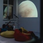 Crianças participam de uma atividade de astronomia com telescópio e projeção de um planeta em ambiente interno.