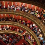 Público acompanha uma apresentação no interior de um teatro histórico, com arquitetura ornamentada e balcões laterais.