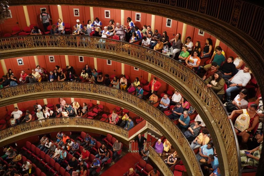 Público acompanha uma apresentação no interior de um teatro histórico, com arquitetura ornamentada e balcões laterais.