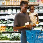 Mulher carrega uma criança no colo enquanto escolhe alimentos na seção de hortifrúti de um supermercado.