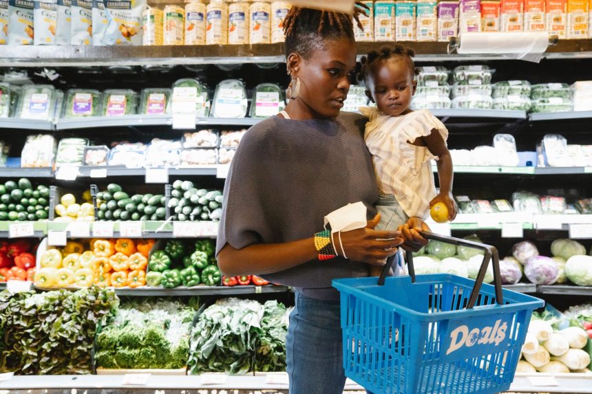 Mulher carrega uma criança no colo enquanto escolhe alimentos na seção de hortifrúti de um supermercado.