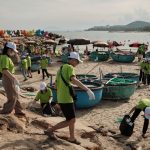 Grupo de voluntários recolhe resíduos na faixa de areia de uma praia durante uma ação de limpeza ambiental.