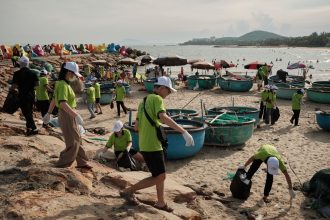 Grupo de voluntários recolhe resíduos na faixa de areia de uma praia durante uma ação de limpeza ambiental.