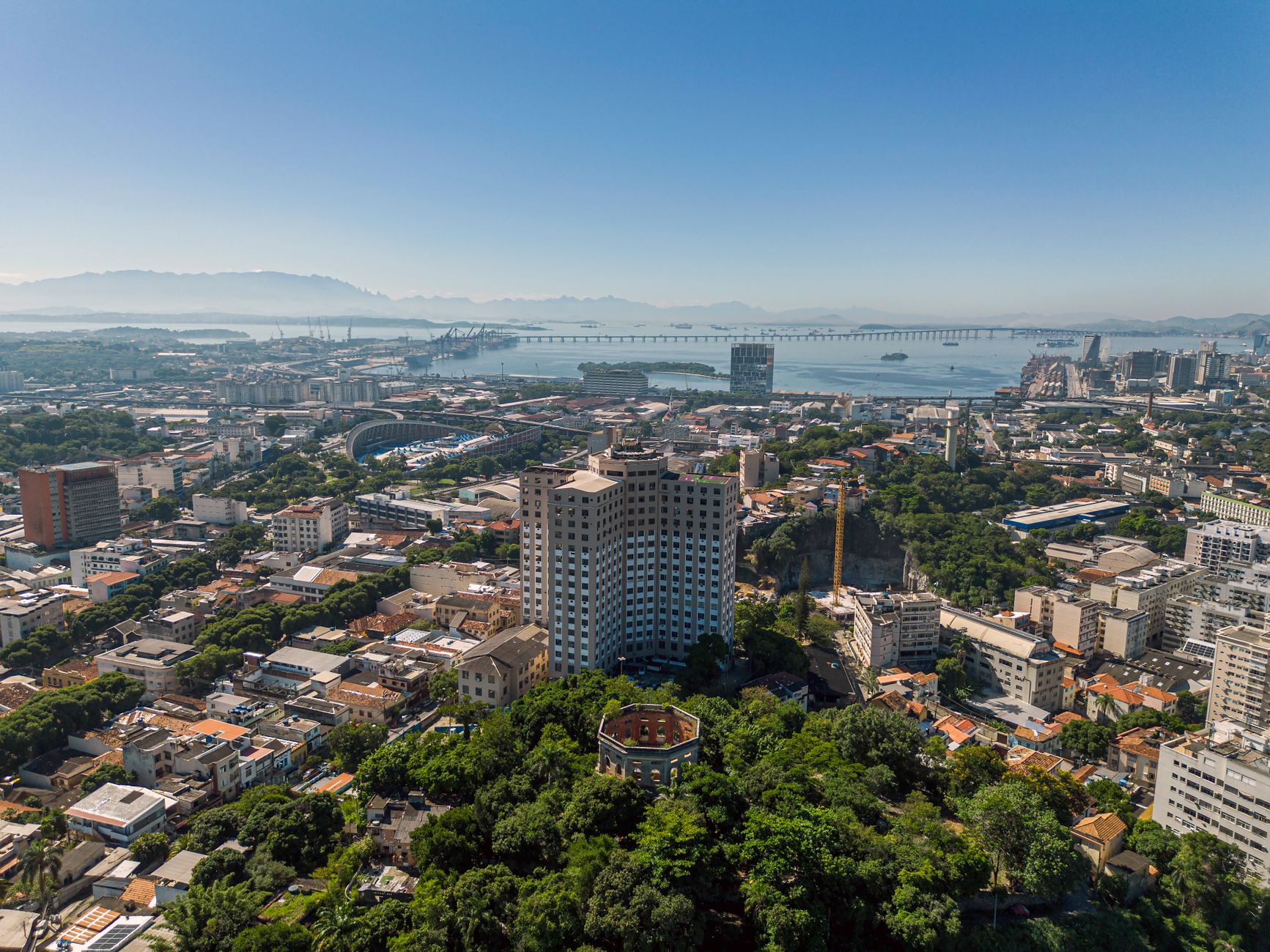 Vista aérea do Rio de Janeiro, com prédios, áreas verdes e o céu azul ao fundo.