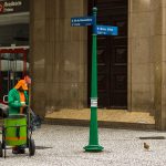 Trabalhador de limpeza urbana atua em calçada de área comercial do Rio de Janeiro, diante de um edifício.