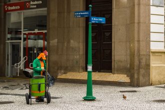 Trabalhador de limpeza urbana atua em calçada de área comercial do Rio de Janeiro, diante de um edifício.