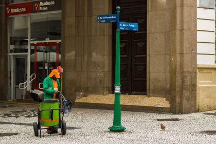 Trabalhador de limpeza urbana atua em calçada de área comercial do Rio de Janeiro, diante de um edifício.