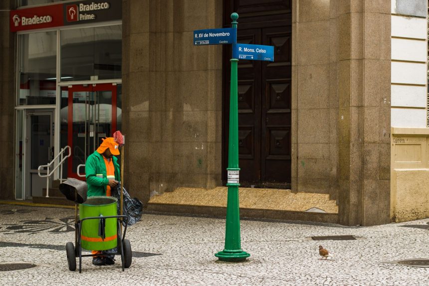 Trabalhador de limpeza urbana atua em calçada de área comercial do Rio de Janeiro, diante de um edifício.
