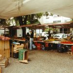 Pessoas circulam por uma feira ao ar livre no Rio de Janeiro, com barracas de produtos frescos sob uma cobertura.