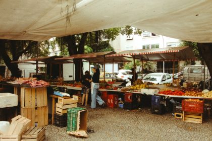 Pessoas circulam por uma feira ao ar livre no Rio de Janeiro, com barracas de produtos frescos sob uma cobertura.
