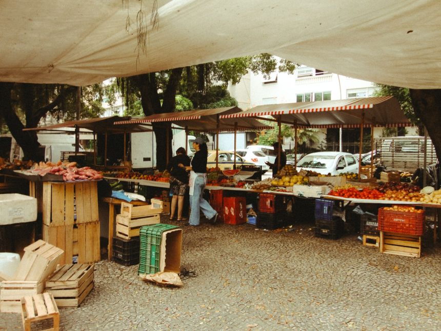 Pessoas circulam por uma feira ao ar livre no Rio de Janeiro, com barracas de produtos frescos sob uma cobertura.